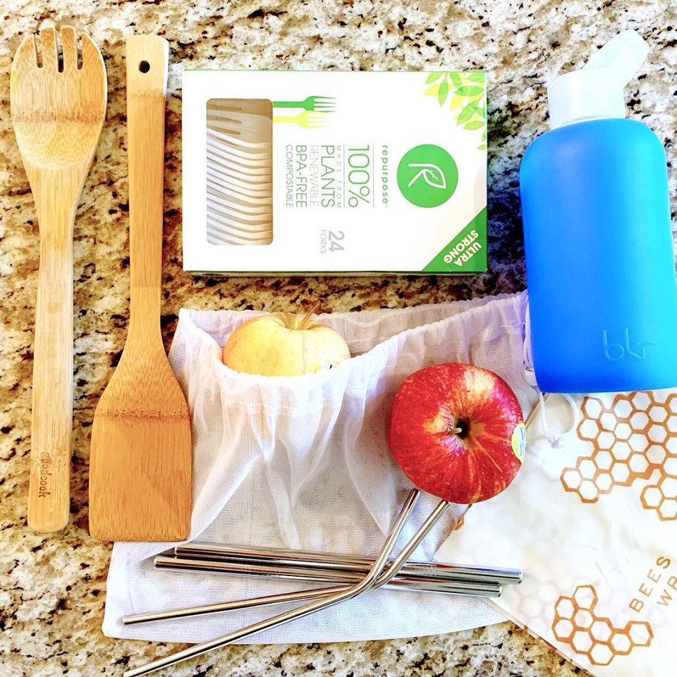 Bamboo spoons, compostable forks, a blue reusable water bottle, apples and a nylon produce bag, beeswax wrap and stainless steel straws on a granite countertop.