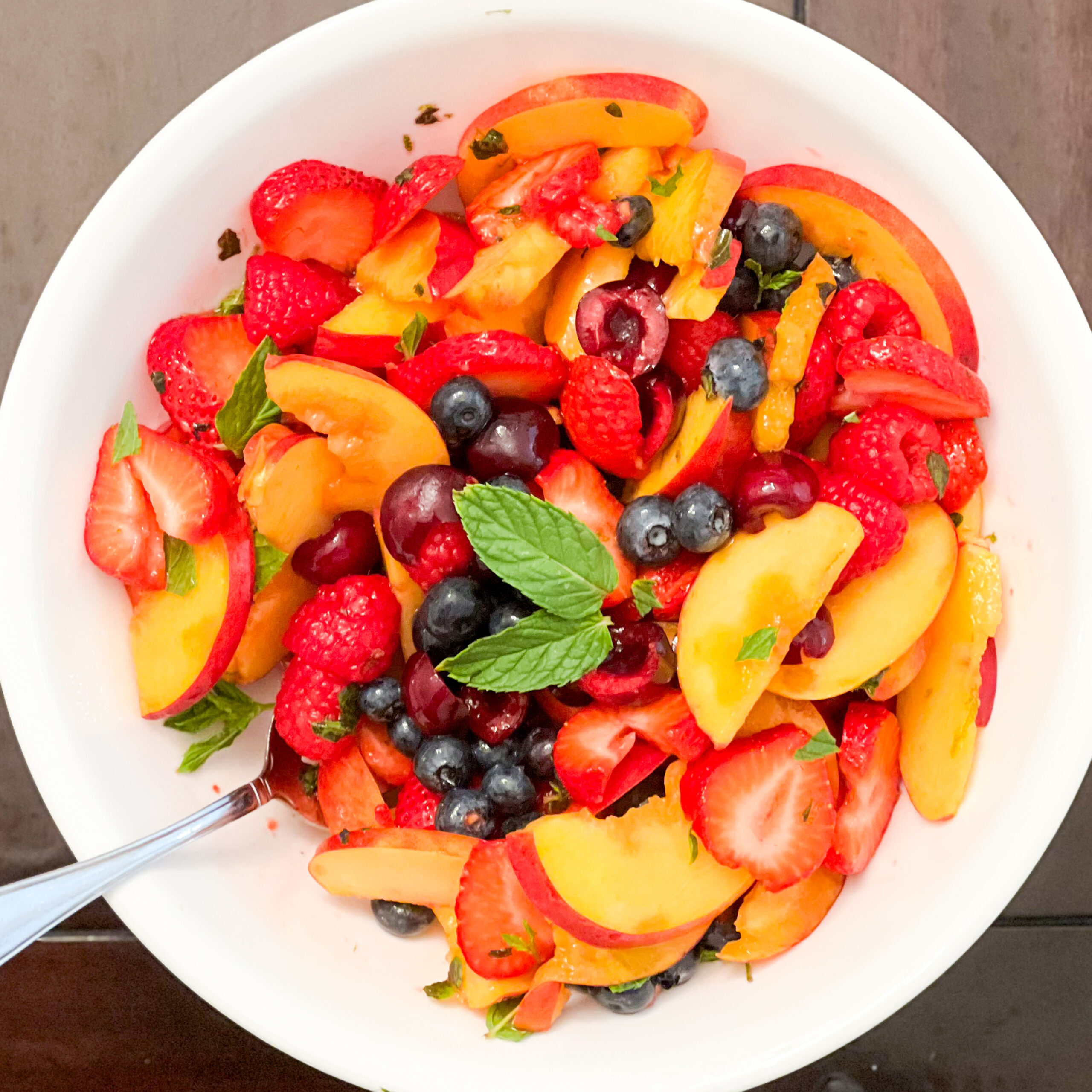 A summer fruit salad in a white bowl with a spoon on a dark brown background.