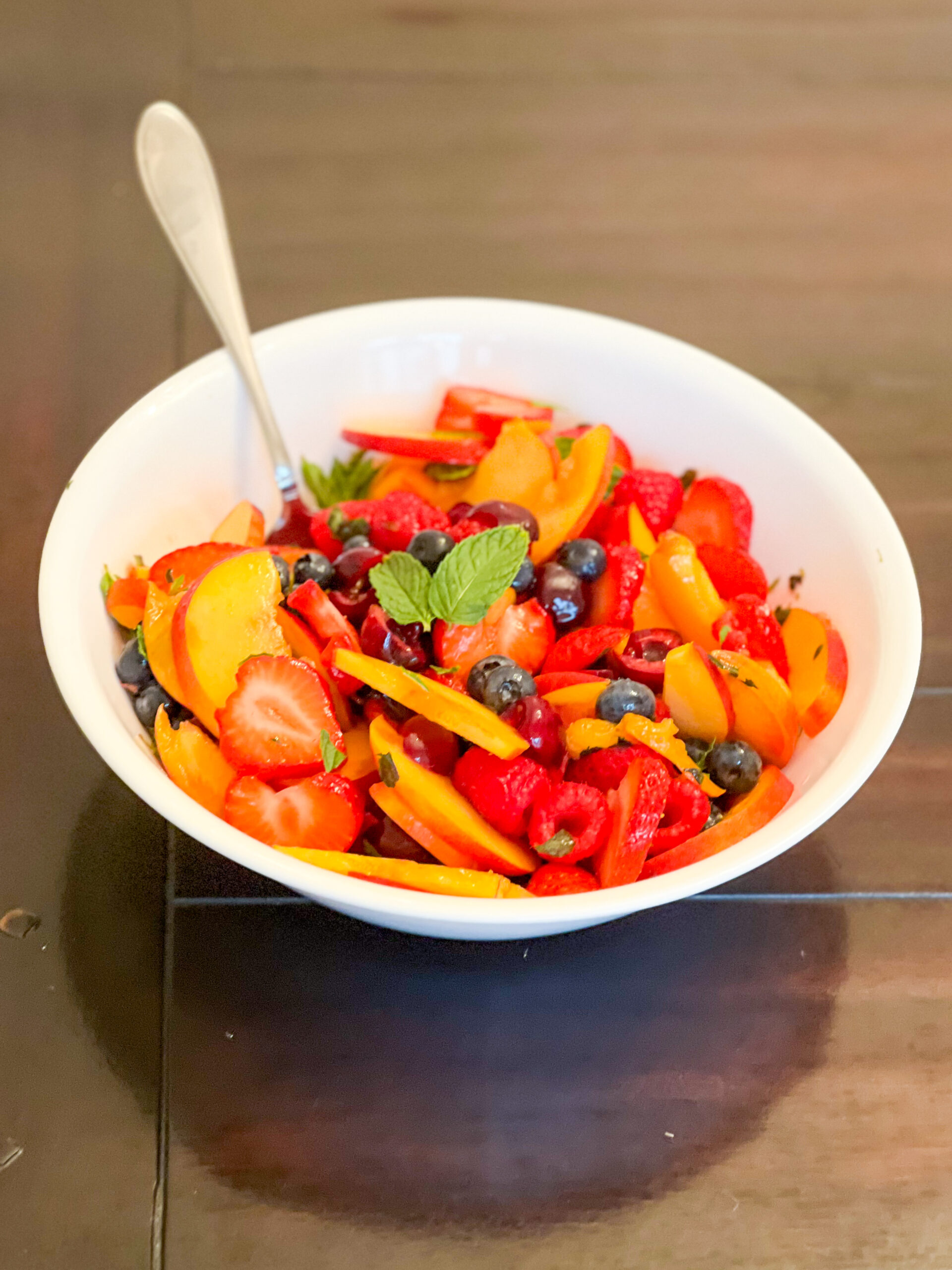 A summer fruit salad in a white bowl with a spoon on a dark brown background.