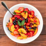 A summer fruit salad in a white bowl with a spoon on a dark brown background.