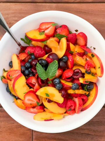 A summer fruit salad in a white bowl with a spoon on a dark brown background.