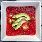 Watermelon beet gazpacho in a white bowl with sliced avocado and tomato.