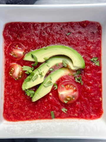 Watermelon beet gazpacho in a white bowl with sliced avocado and tomato.