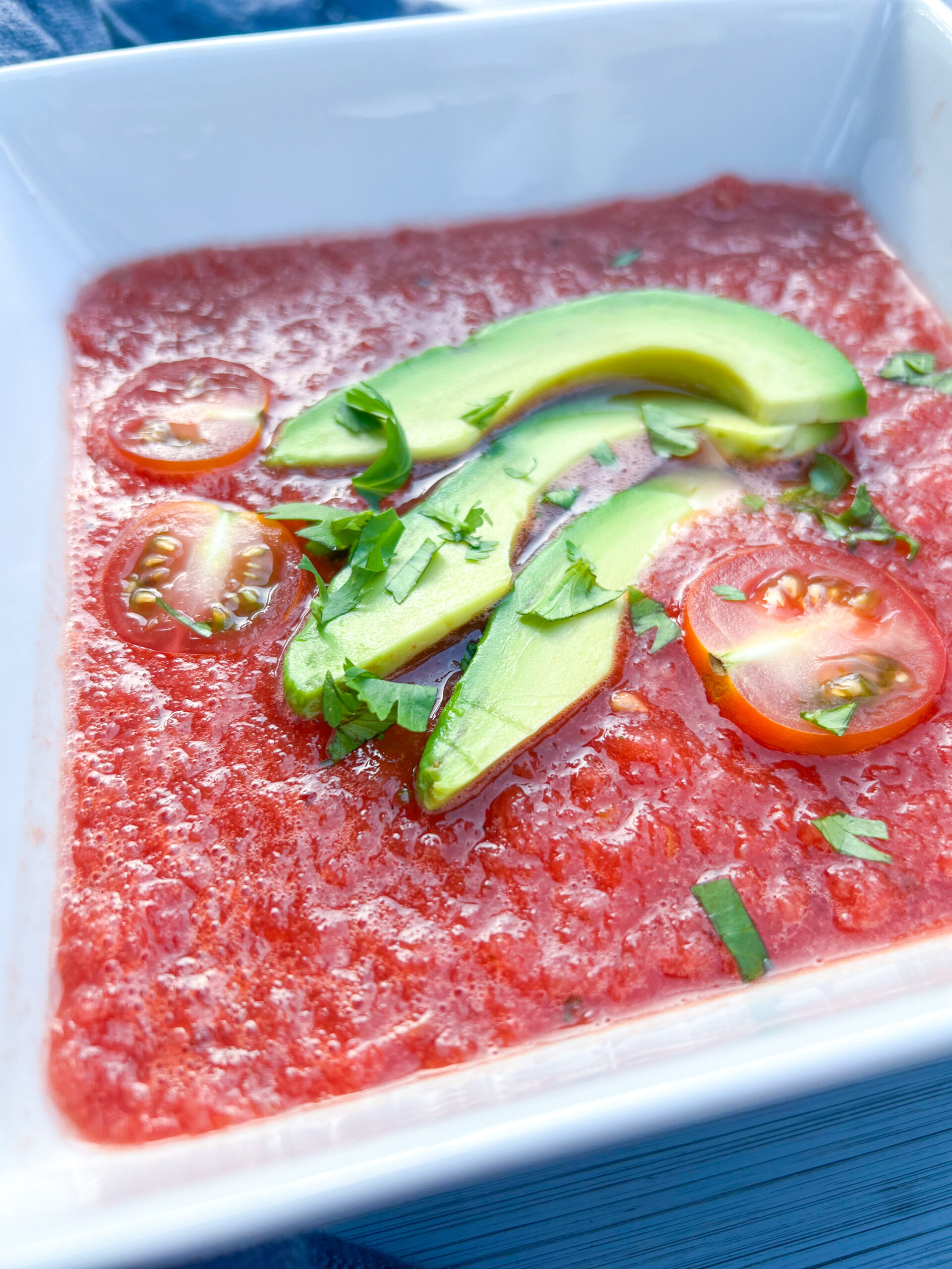 Watermelon beet gazpacho in a white bowl with sliced avocado and tomato.