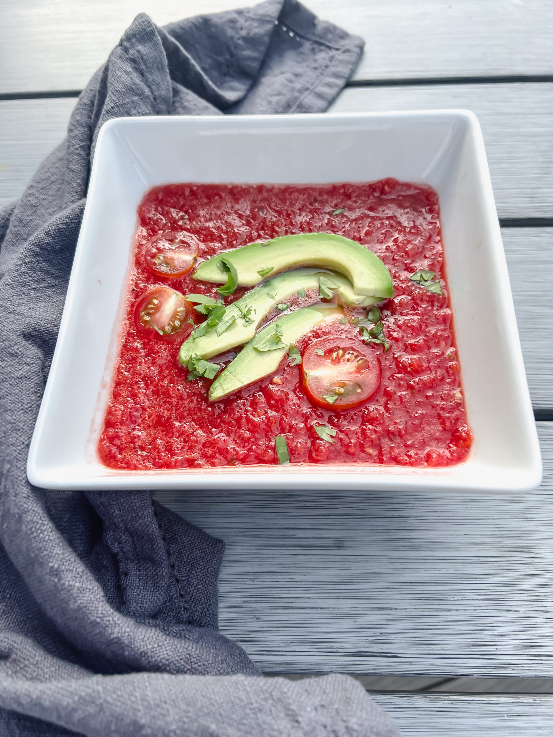 Watermelon beet gazpacho in a white bowl on a picnic table with sliced avocado and tomato and a gray napkin on the side.