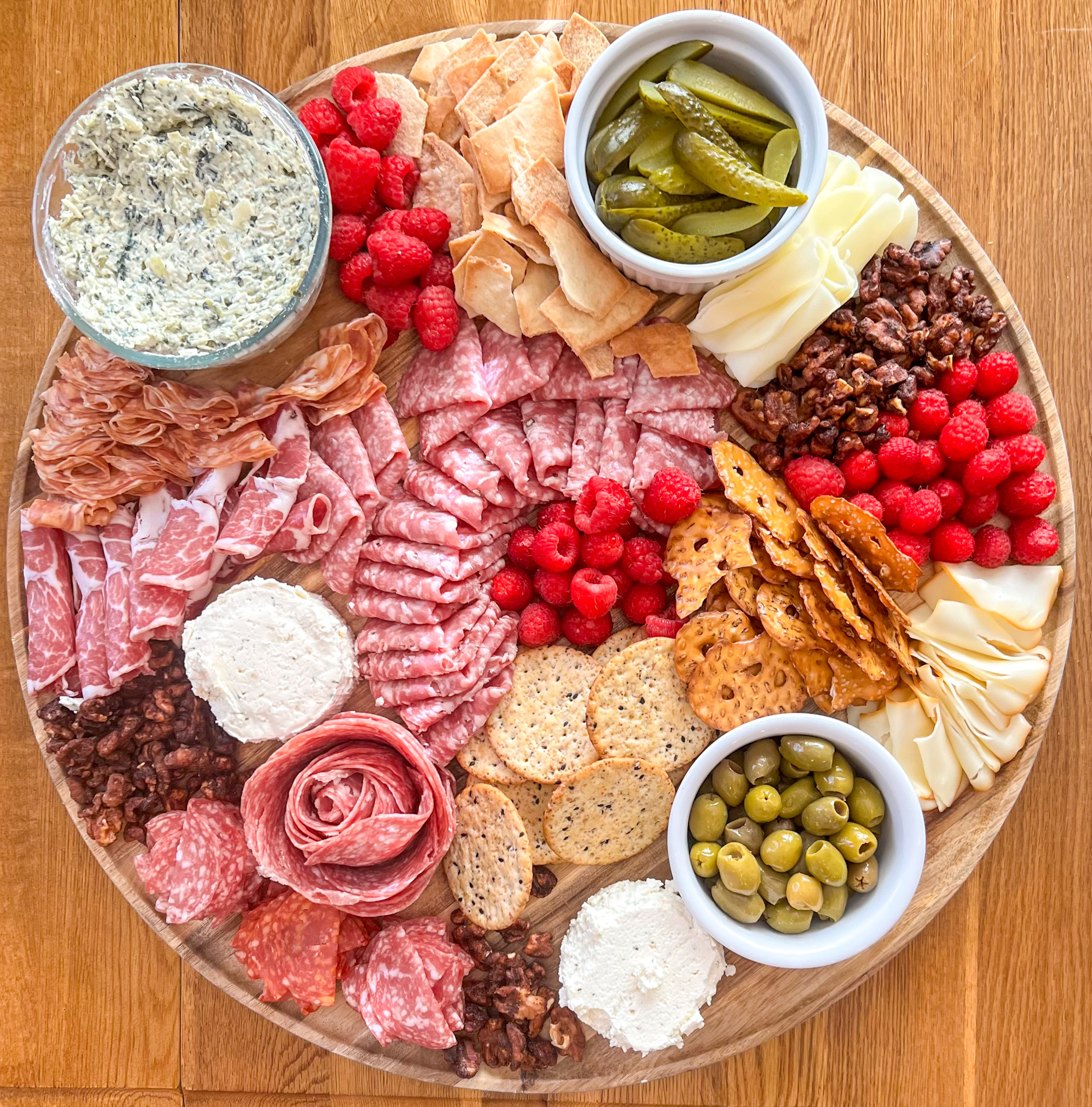 A styled charcuterie board wwith meat, cheese, dips, crackers, olives, pickles, nuts, and raspberries on a round wooden board.