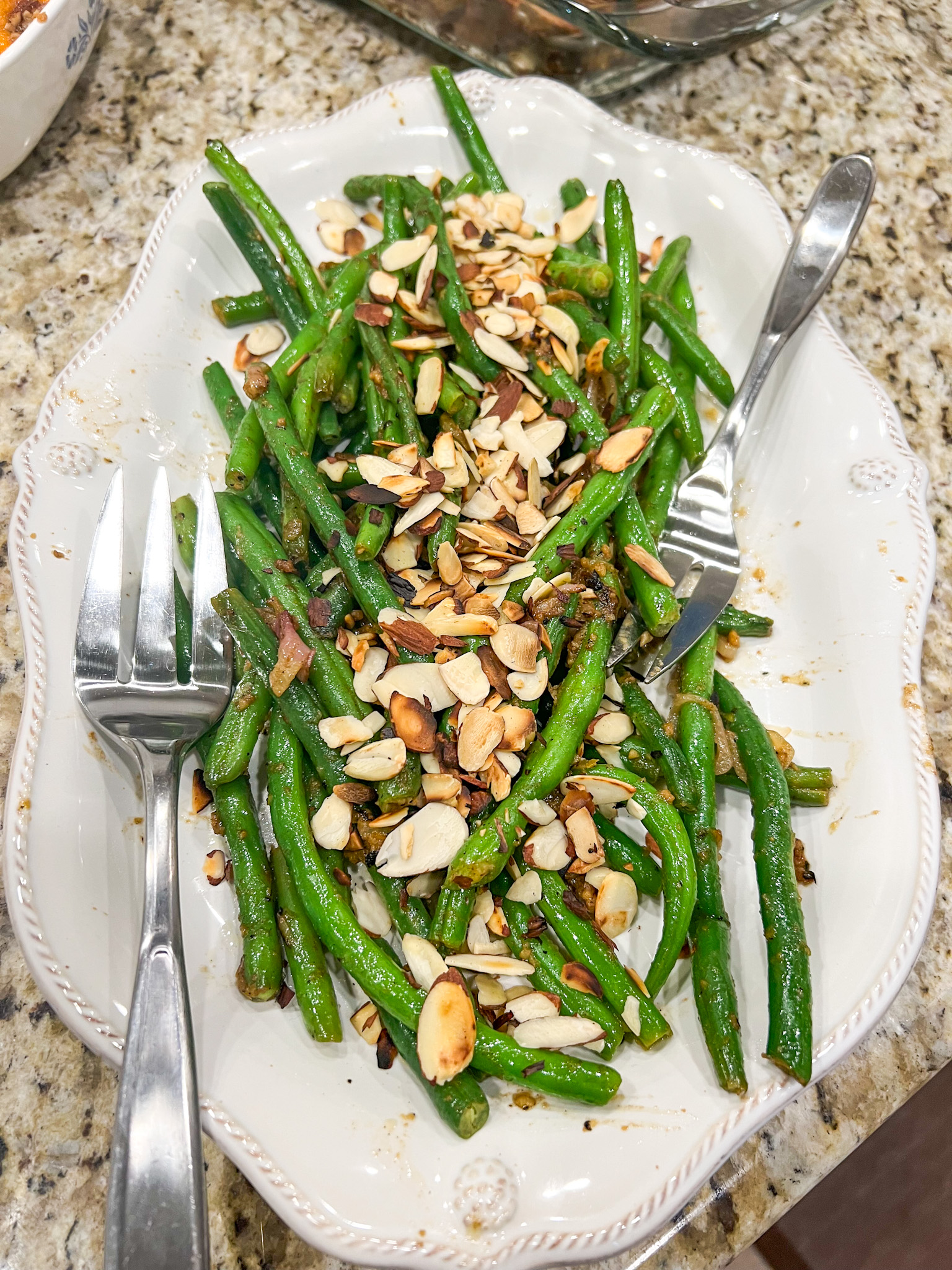 Sautéed green beans with almonds and a garlic lemon sauce on a white serving dish with silver serving utensils.