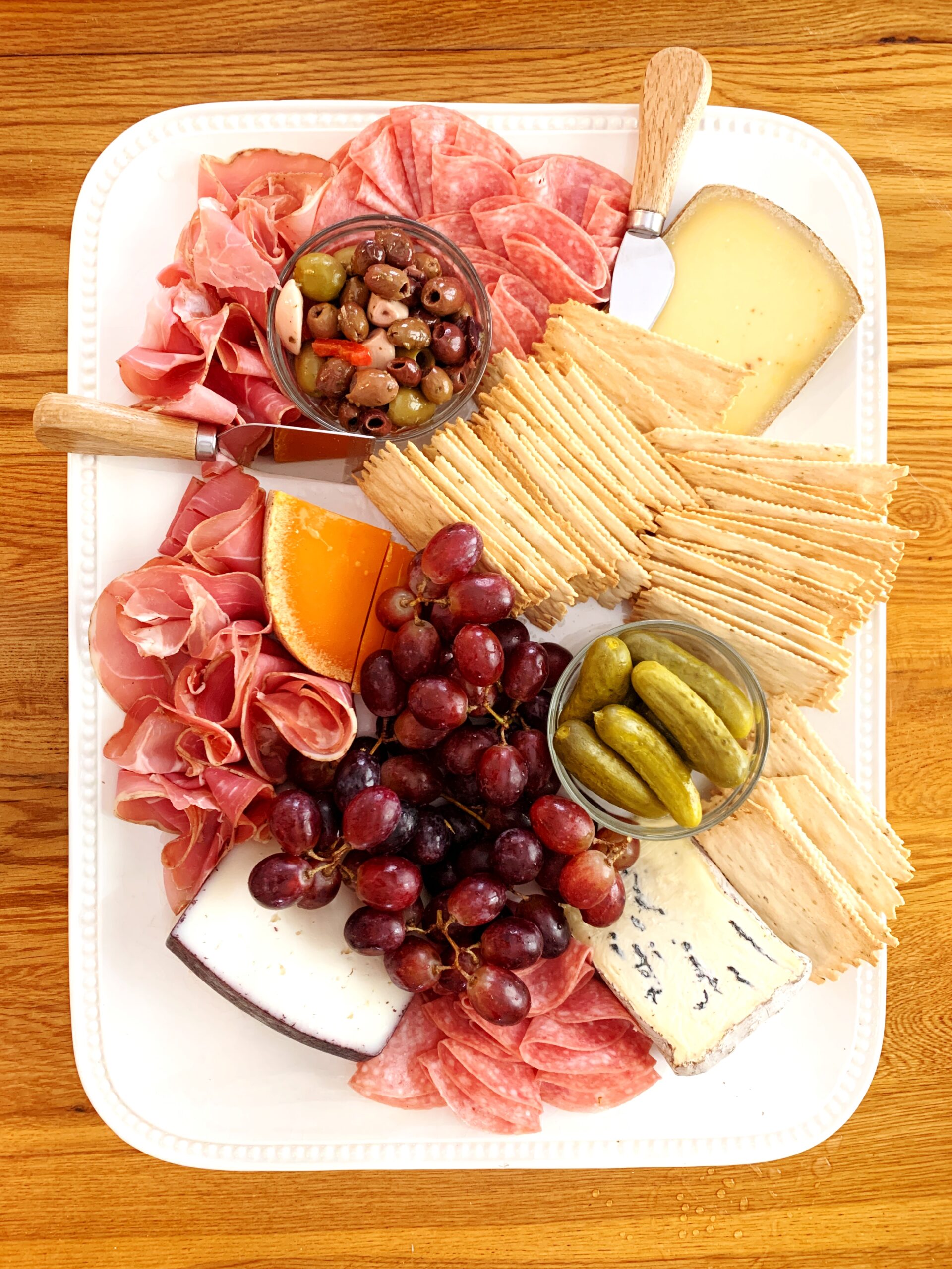 A charcuterie board with meat, cheese, crackers, pickles, olives and grapes on a white platter on a wood table