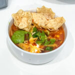 Beef chili in a bowl on a white countertop with toppings of shredded cheese, sour cream, cilantro, avocado and tortilla chips.