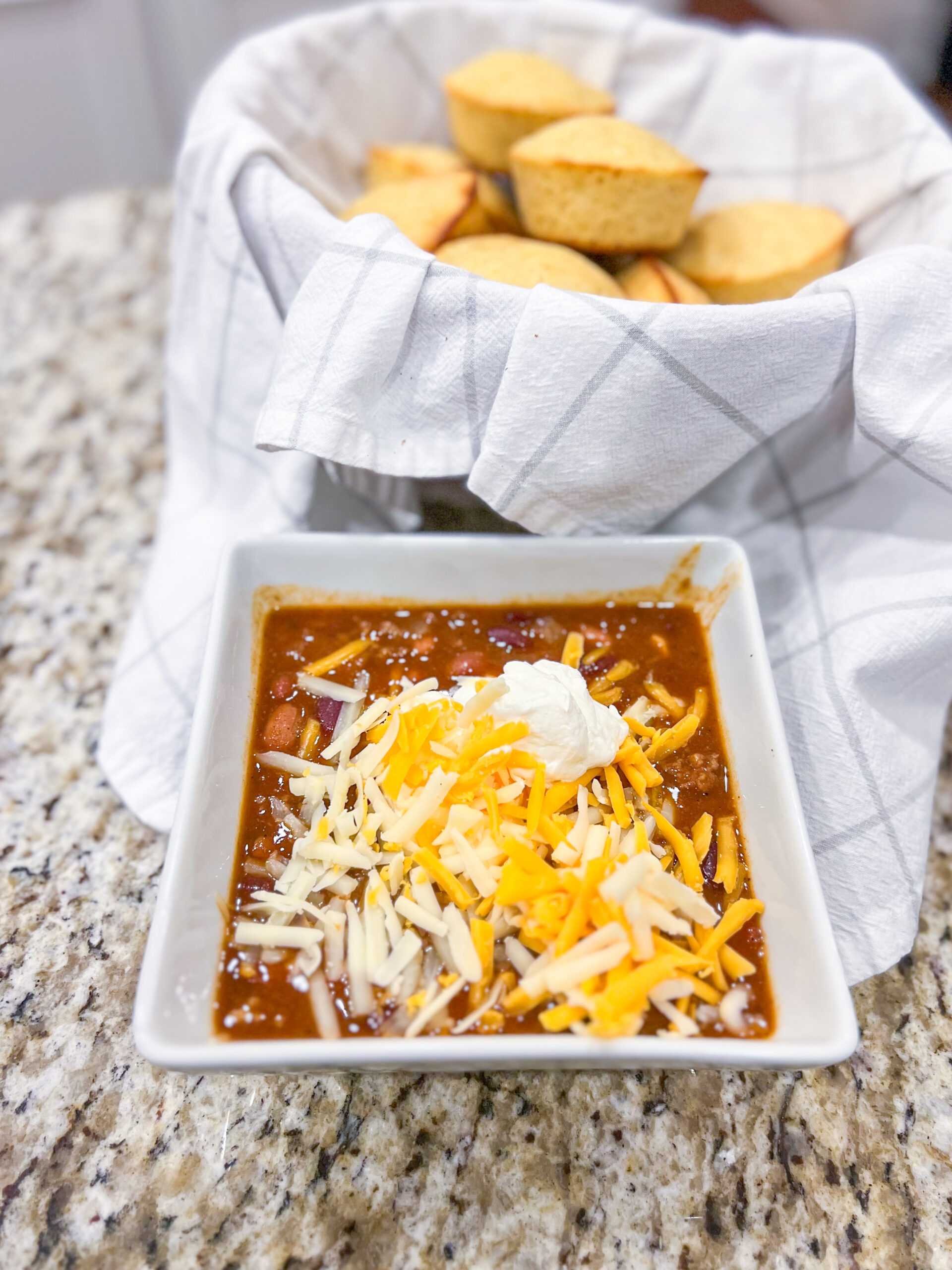 Beef chili in a white bowl with shredded cheese and sour cream on top with a basket of cornbread muffins in the background.